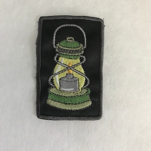May include: A black and green embroidered patch of a lantern with a yellow flame.