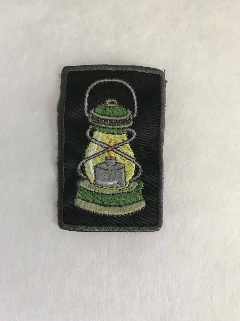 May include: A black and green embroidered patch of a lantern with a yellow flame.