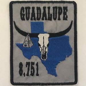 May include: Embroidered patch with the word "GUADALUPE" above a blue Texas outline. Features a steer skull, horns, and a cheese wedge. The number "8,751" is below the Texas outline. Gray background with black border.