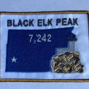 Puede incluir: Parche bordado blanco con borde dorado. El parche presenta un contorno azul de una montaña con una estrella blanca, el texto "BLACK ELK PEAK" y el número "7,242".