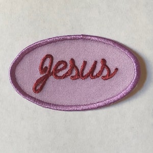 Pode incluir: Patch bordado oval em tons de roxo e rosa. A palavra "Jesus" é costurada em fonte cursiva, bordô, no centro. O patch tem um acabamento acetinado e uma borda costurada.