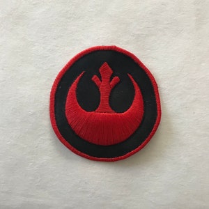May include: A red embroidered patch with a black outline, featuring the Rebel Alliance symbol from Star Wars.