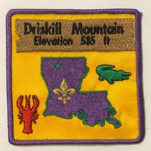Puede incluir: Un parche bordado amarillo con un contorno morado. El parche presenta un mapa de Luisiana con una flor de lis, un caimán verde y un cangrejo rojo. El texto "Driskell Mountain Elevation 585 ft" está en una pancarta marrón en la parte superior.