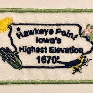 Puede incluir: Parche bordado con el texto "Hawkeye Point, Iowa's Highest Elevation 1670'". El parche presenta el contorno de Iowa, maíz bordado, un pájaro y flores, con un borde verde. La elevación es de 509 metros.