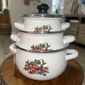 May include: A stack of three white enamel pots with lids and handles. Each pot features a colorful fruit and vegetable design. The pots are stacked on a wooden surface, tied together with twine.