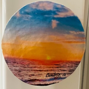 Può includere: Un adesivo rotondo con un tramonto sull'oceano. Il cielo è un gradiente di blu, rosa e arancione. Il sole tramonta in lontananza e l'oceano è di un blu profondo. L'adesivo dice "Cambria, CA" nell'angolo in basso a destra.