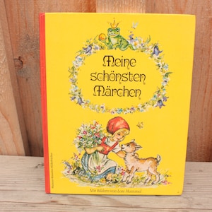 Könnte beinhalten: Ein gelbes Buch mit rotem Rücken, betitelt "Meine schönsten Märchen", mit einem Bild eines Mädchens in einer roten Haube, das einen Blumenstrauß und eine kleine Ziege hält. Das Buch hat einen floralen Rahmen mit einem Frosch, der eine Krone trägt.