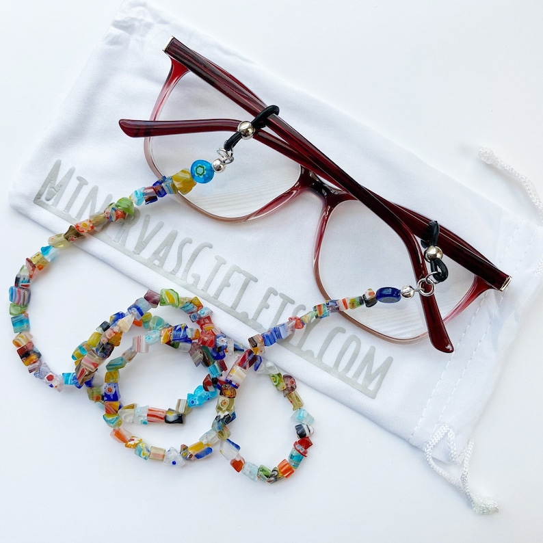 The picture shows a colorful array of cube shaped chip beads strung with adjustable loops that attach to a pair of reading glasses. The glasses rest on a white storage pouch with the name of the shop printed on it.