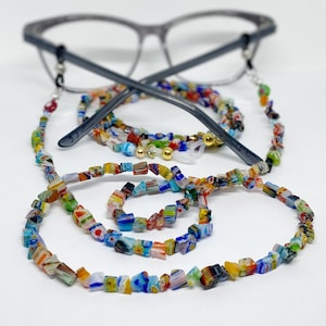 A beaded strand with a rainbow of colorful, floral patterned chip beads sits in front of a pair of reading glasses.