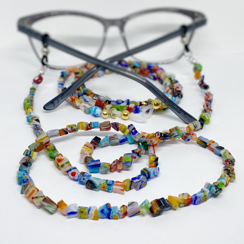 A beaded strand with a rainbow of colorful, floral patterned chip beads sits in front of a pair of reading glasses.