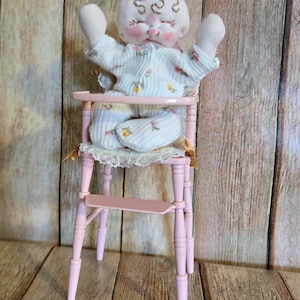 May include: A handmade doll in a pink high chair. The doll is wearing a white floral patterned outfit and has its arms raised. The pink high chair has a lace seat and a footrest. The background is a wooden wall.