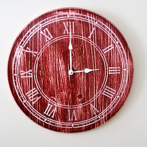 Puede incluir: Reloj de pared redondo rojo con números romanos y manecillas blancas. La esfera del reloj tiene una textura de vetas de madera. Las manecillas señalan aproximadamente las 3 en punto. Un elemento decorativo para el hogar.