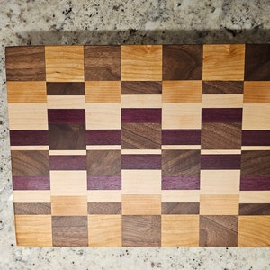 May include: A wooden cutting board with a checkerboard pattern made from walnut, maple, and purpleheart wood.