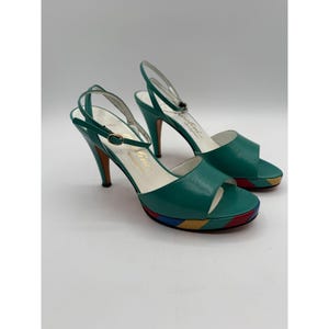 May include: A pair of teal green high-heeled sandals. The sandals have a platform sole with red, yellow, and blue stripes. The heels are a curved design with a brown finish. The ankle straps are silver. The sandals are labeled "Caroline".