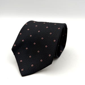 Op de afbeelding: Een zwarte das met een herhalend patroon van kleine, lichtgekleurde ruitvormen, elk met een minuscuul roze paisley-motief. De achtergrondstof is getextureerd met een subtiel, fijn geometrisch patroon van korte, dunne, donkergrijze lijntjes.