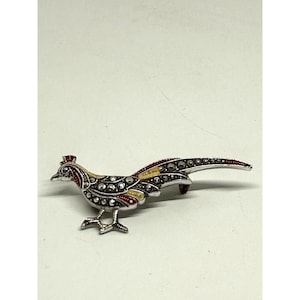 Puede incluir: Broche de pájaro plateado con un diseño detallado. El broche presenta un faisán con detalles rojos, amarillos y de cristal transparente. La cola del pájaro es larga y curvada, y el broche tiene un cierre de alfiler en la parte posterior.