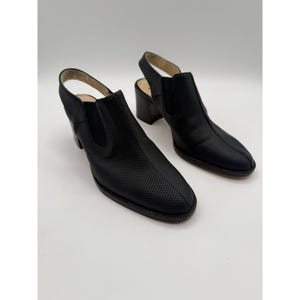 Puede incluir: Un par de zapatos negros con tacón abierto y textura perforada en la pala. Los zapatos tienen punta redondeada, tacón pequeño y una correa que rodea el tobillo.