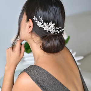 以下が含まれることがあります： 花柄のデザインとクリスタルをあしらったシルバーのヘアコーム。女性の髪につけられています。