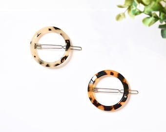May include: Two tortoise shell hair clips with a round shape and a metal bar in the center. The clips are on a white background.