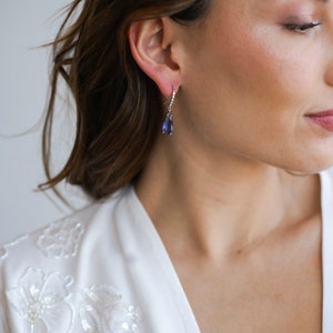 May include: A woman wearing a white top with floral embroidery and a pair of silver teardrop earrings with a purple gemstone.