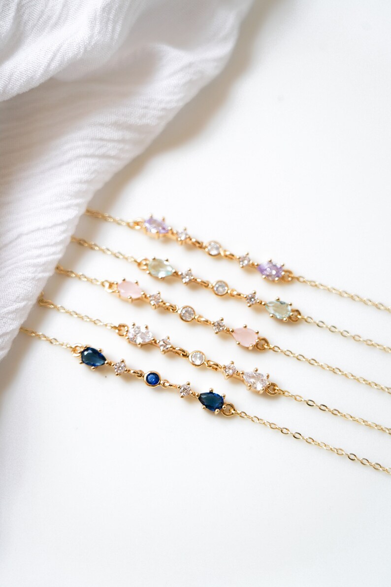 May include: Five gold chain bracelets with different colored gemstones. Each bracelet features a delicate chain with small clear stones and larger pear-shaped stones in shades of blue, pink, and green.