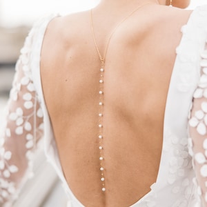 Puede incluir: Un vestido de novia blanco con la espalda baja y un detalle de cadena de oro y perlas. El vestido tiene una superposición de encaje con diseños florales.