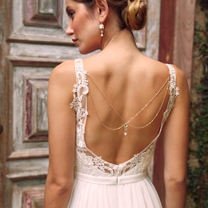 Puede incluir: Un vestido de novia blanco con un corpiño de encaje y una falda fluida. El vestido presenta una espalda baja con una delicada cadena dorada y un detalle de perla.