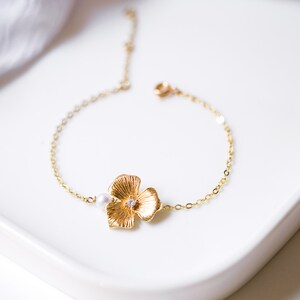 May include: A gold chain bracelet with a delicate flower charm and a single pearl accent. The bracelet is on a white surface.
