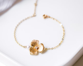 May include: A gold chain bracelet with a delicate flower charm and a single pearl accent. The bracelet is on a white surface.
