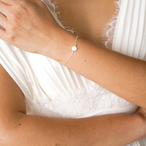 May include: A delicate silver chain bracelet with a single white oval charm. The bracelet is worn on a person's wrist.