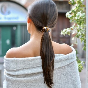 Può includere: Una donna con lunghi capelli castani raccolti in una coda di cavallo con un elastico per capelli marrone e nero a tartaruga. Indossa un maglione grigio chiaro a spalle scoperte.