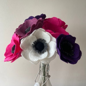 Może przedstawiać: Bukiet filcowych kwiatów anemonów w przezroczystej szklanej butelce. Kwiaty są w odcieniach różu, fioletu i bieli, z ciemnoszarymi środkami. Butelka ma metalowe i plastikowe zamknięcie.