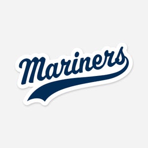 May include: A die-cut sticker featuring the word "Mariners" in a navy blue script font with a white border. The word has a stylized tail that curves beneath the text.