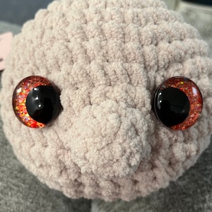May include: Close-up of a plush toy's face with red glitter eyes. The toy is made of a soft, light pink material.
