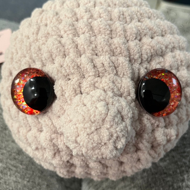 May include: Close-up of a plush toy's face with red glitter eyes. The toy is made of a soft, light pink material.