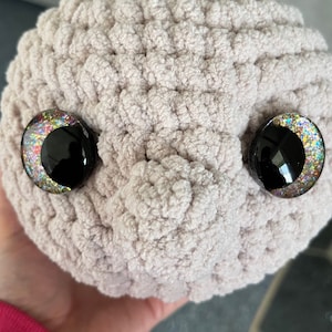 May include: A close-up of a crocheted plush toy with large, glittery eyes. The toy is light brown and has a textured surface.
