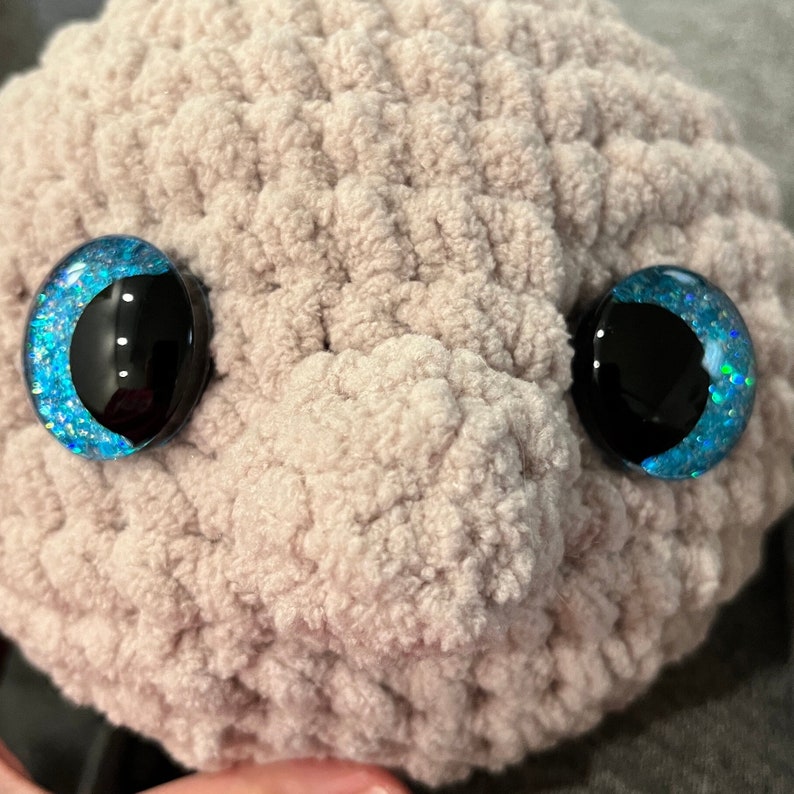 May include: Close-up of a plush toy's face with large, blue, glittery eyes. The toy is made of a soft, beige, fuzzy material.