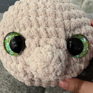 May include: A close-up of a crocheted plush toy in a light brown colour. The toy has two large, round eyes with black pupils and green, blue and gold glitter accents.