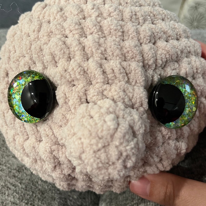 May include: A close-up of a crocheted plush toy in a light brown colour. The toy has two large, round eyes with black pupils and green, blue and gold glitter accents.