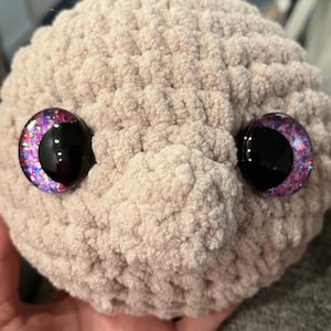 May include: A close-up of a crocheted plush toy with large, glittery eyes. The eyes are purple, pink, and black. The toy is made of a soft, fuzzy material.