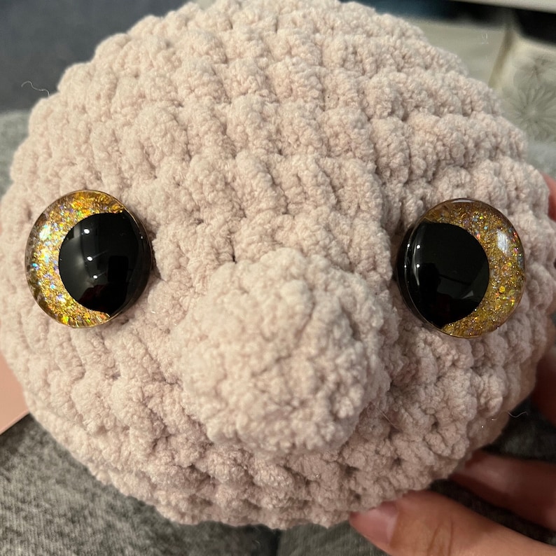 May include: A plush, light pink, crocheted toy with a round shape and two large, black eyes with gold glitter accents.