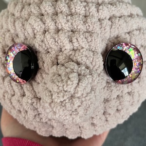May include: A close-up of a plush toy with pink and iridescent glitter eyes. The toy is made of a soft, fuzzy material and is light brown in colour.