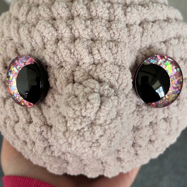 May include: A close-up of a plush toy with pink and iridescent glitter eyes. The toy is made of a soft, fuzzy material and is light brown in colour.