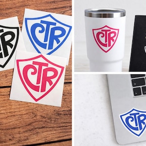 Puede incluir: La imagen muestra varios artículos con un diseño de escudo "CTR". Incluye pegatinas en negro, azul y rosa, un vaso blanco, una Biblia negra y una pegatina para portátil. Los artículos se muestran sobre una superficie de madera y un portátil.