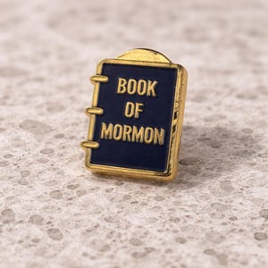 Könnte beinhalten: Ein goldfarbener Ansteckpin in Buchform mit den Worten "BOOK OF MORMON" in goldener Schrift auf marineblauem Hintergrund. Der Pin hat eine rechteckige Form mit abgerundeten Ecken und einem kleinen Verschluss.
