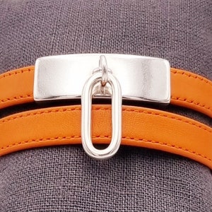 Op de afbeelding: Een oranje leren armband met dubbele wikkeling, een rechthoekige zilveren plaat en een ovale zilveren hanger.