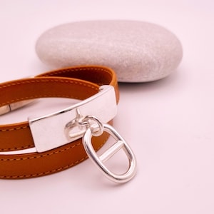 Op de afbeelding: Een bruin leren armband met een zilveren bedel. De bedel is een grote ovaal met een kleinere ovaal erin.