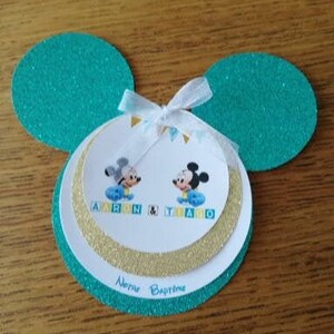 Peut inclure: Une carte en forme de Mickey Mouse bleu turquoise avec des accents de paillettes dorées. La carte représente deux personnages de dessin animé de Mickey Mouse avec le texte "Aaron & Tiago" et "Novas Baptisme".