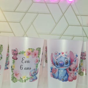 May include: Clear plastic cups decorated with Stitch and Angel characters. One cup has the text "Eva 6 ans" surrounded by flowers. A neon sign in the background displays "good vibes".