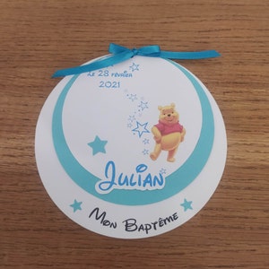 Peut inclure: Une carte circulaire blanche et bleue avec un ruban bleu. La carte représente un personnage de Winnie l'ourson, des étoiles et le texte "Le 28 Février 2021", "Julian" et "Mon Baptême".